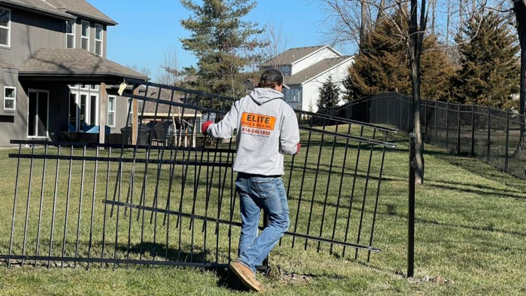 Elite Fence installer carrying a black ornamental fence panel during installation in a residential neighborhood in Raymore MO.