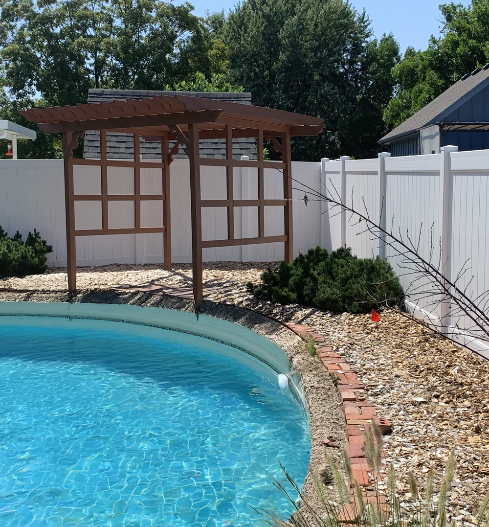A white vinyl pool safety fence with custom wooden arbor gate and semi-private panels surrounding a curved swimming pool.