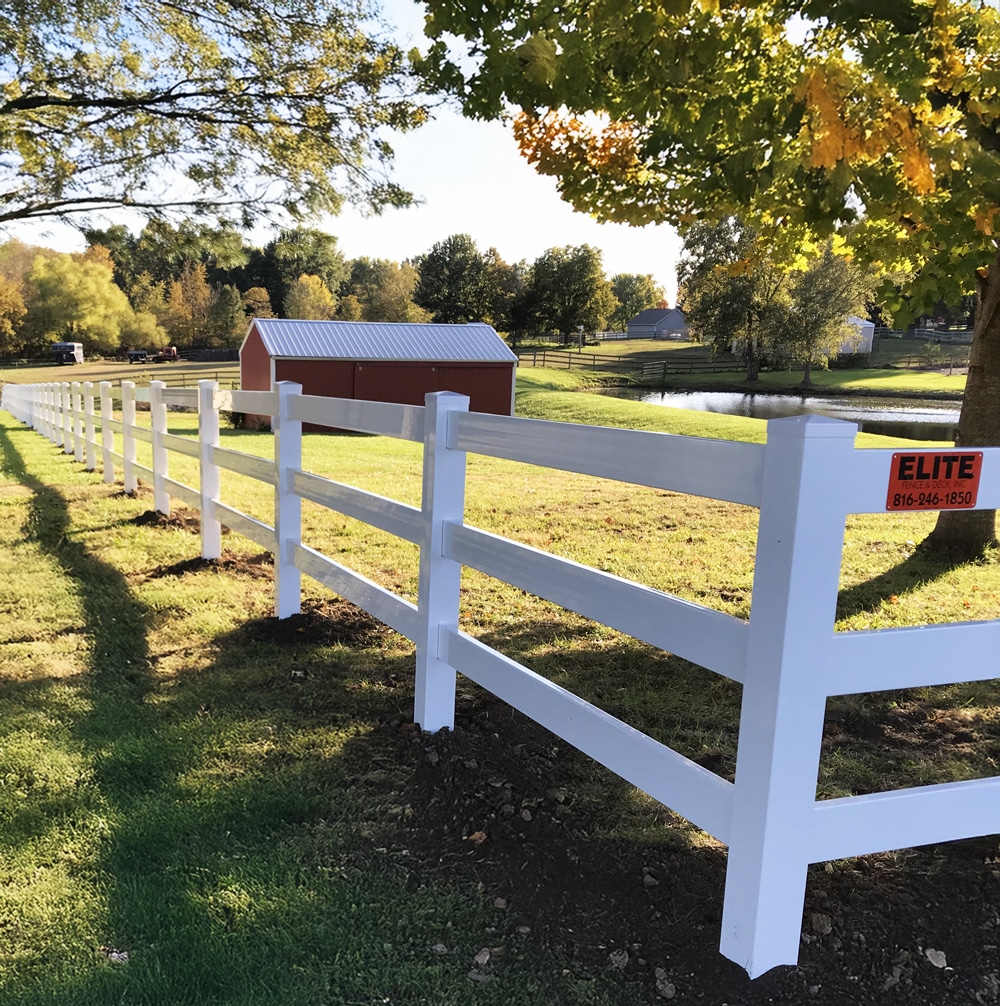 Elite Fence installation of a white vinyl post and three-rail ranch fence bordering a grassy field and pond.