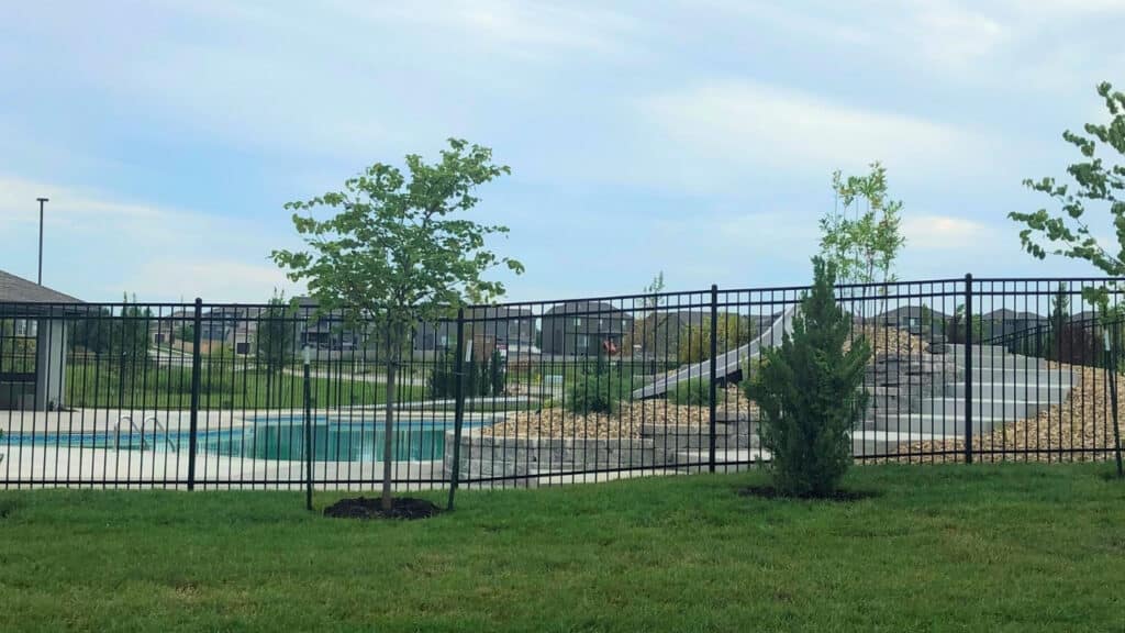 Black aluminum pool safety fence installed around a residential backyard swimming pool with a stone retaining wall and slide.
