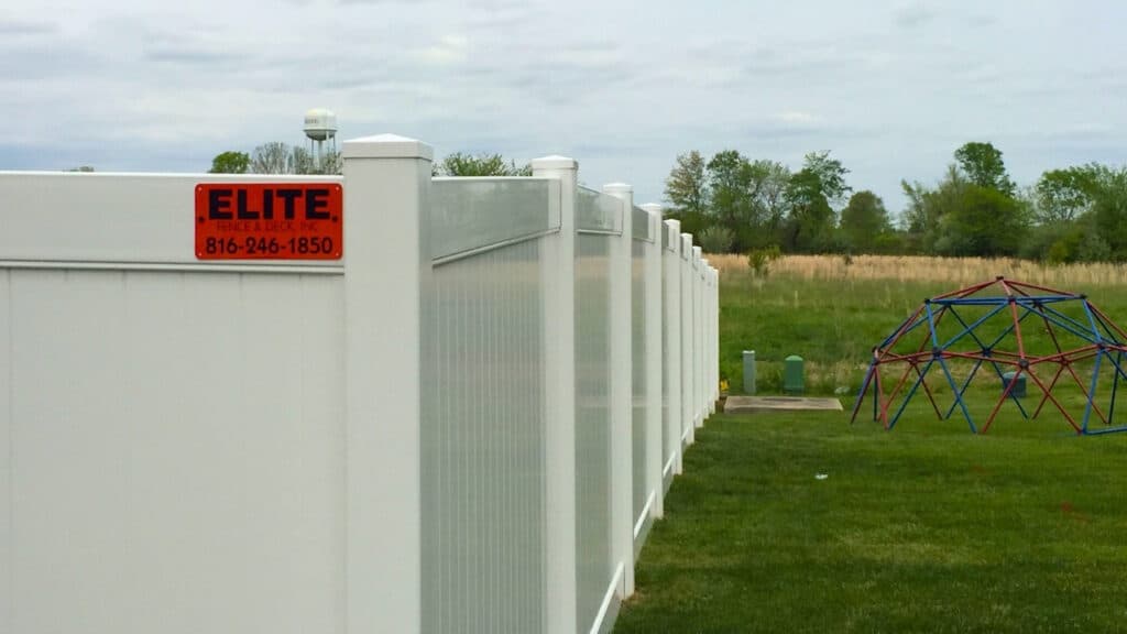 A long white vinyl privacy fence installed in a grassy backyard with an Elite Fence & Deck sign, demonstrating durable vinyl fencing.