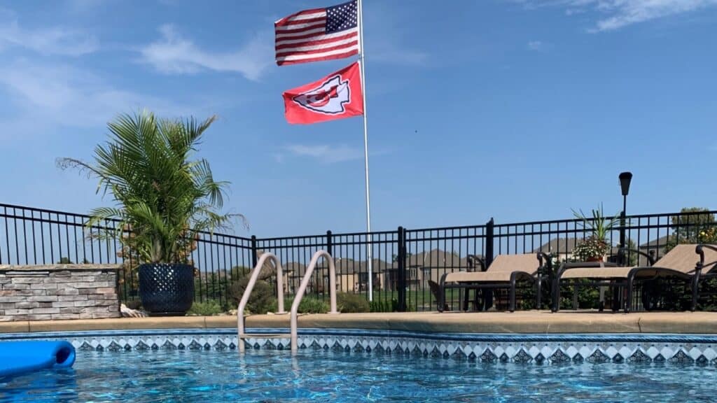Black aluminum pool safety fence surrounding a residential swimming pool with lounge chairs and a Kansas City Chiefs flag.
