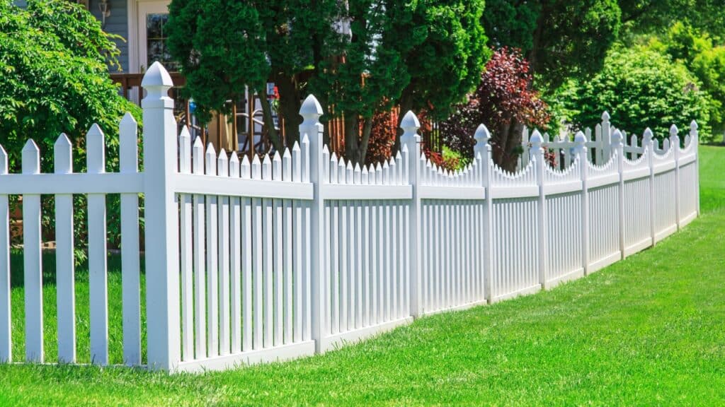 White vinyl picket fence with decorative gothic post caps installed along a green front lawn, highlighting front garden fence ideas.