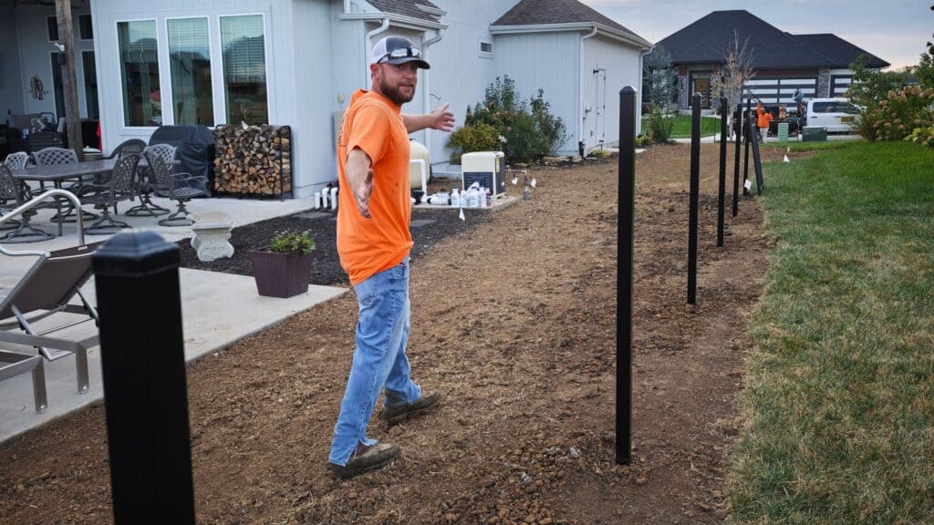 Elite Fence installer gesturing along a newly dug line of set black fence posts during a residential yard fence replacement.