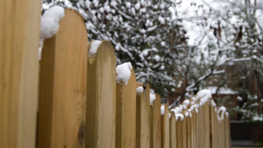 Snow resting on the pickets of a newly installed wooden privacy fence during winter.